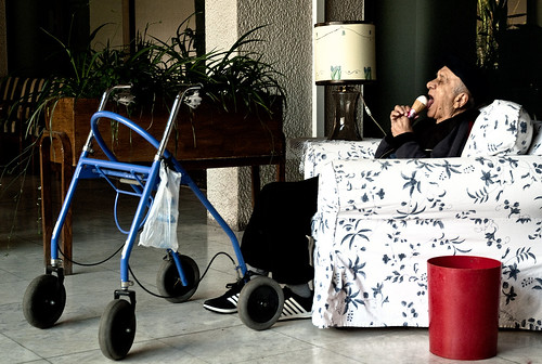 an elderly man sitting in a white chair with floral print, eating a vanilla ice cream cone. There is a red trashcan next to him, empty. In front of him is his walker.